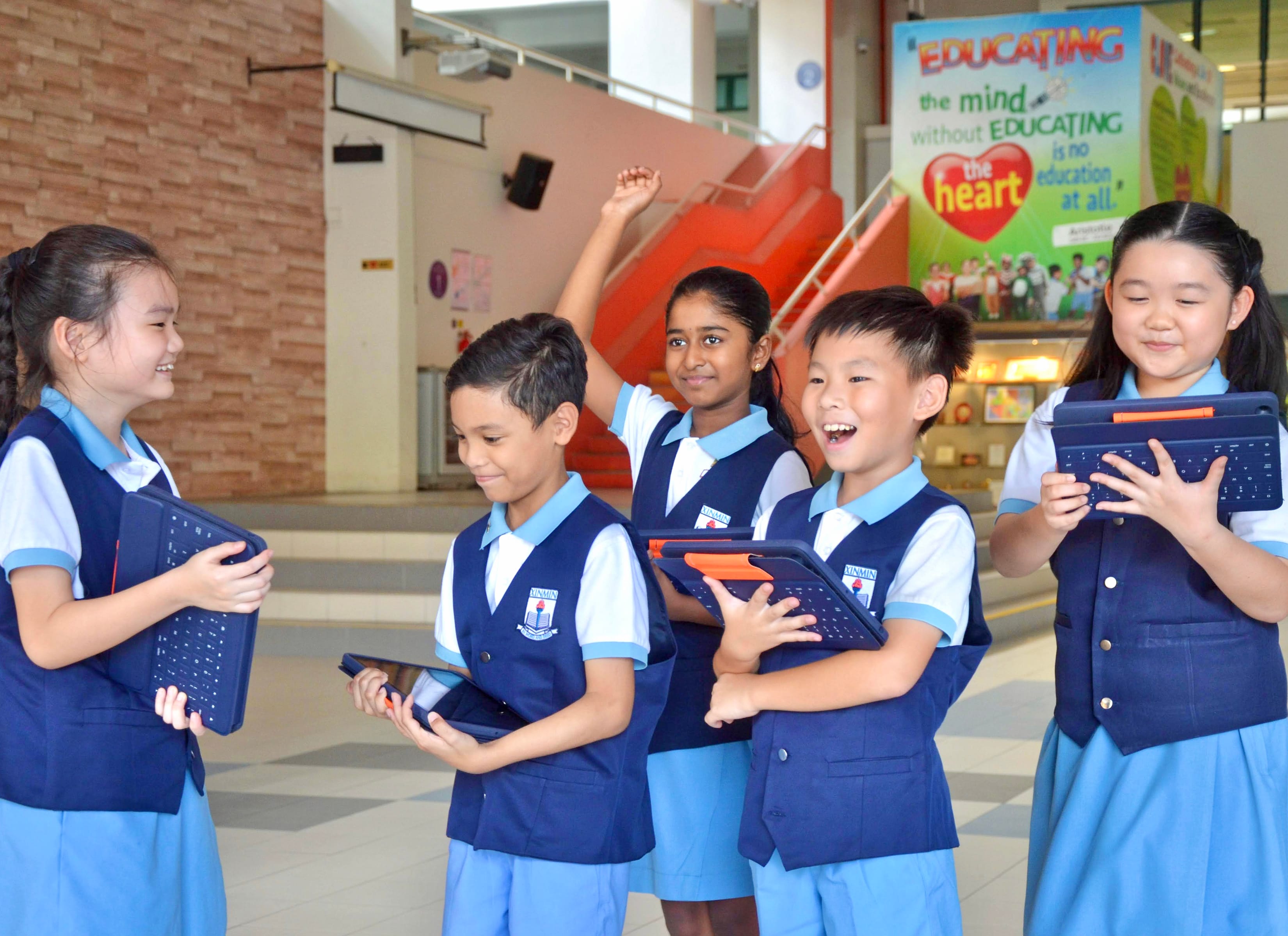 Five smiling children in blue school uniforms hold tablets with keyboards inside a school building.